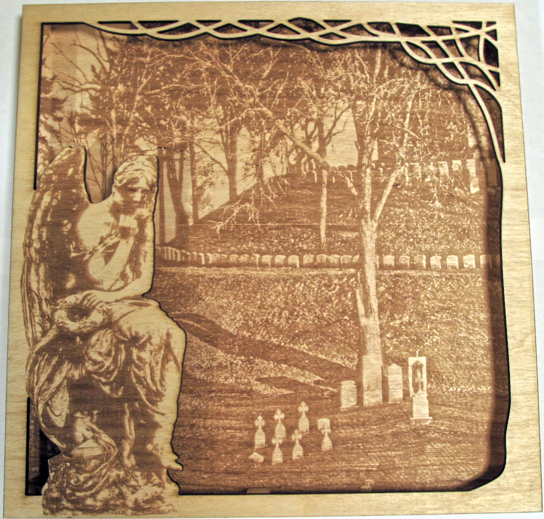 This is a laser cut wall hanging. The back piece is an image of a hill in a graveyard and the top piece is a part of a tree with an angel statue on the opposite side.